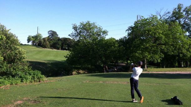Golfer teeing off toward a tree-lined fairway at Hancock Golf Course in Austin.